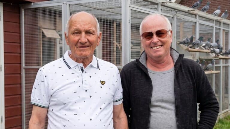 A cheerful photo of pigeon fanciers Geert Hoezen and Wim Steinhage standing together outside their racing loft in Belfeld, smiling proudly after their spectacular double provincial victory.