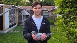 A close-up photograph of young Belgian fancier Milan van Win smiling warmly at the camera while gently holding a champion racing pigeon in both hands. He is wearing a dark casual jacket and standing outdoors in front of a traditional wooden pigeon loft with open landing boards. The pigeon is a beautifully groomed blue bar racing pigeon with clear markings, looking alert and calm. The lighting is natural and bright, capturing a proud, authentic moment between the fancier and his bird.