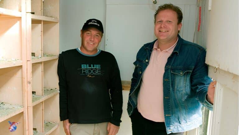 Johan van de Kimmenade and Dirk Verbakel, the pigeon racing duo from Helmond, smiling brightly inside their wooden pigeon loft following their spectacular victory. On the left, one partner wears a black baseball cap, a black long-sleeve shirt with a Blue Ridge logo, and light-colored trousers. On the right, his partner smiles warmly in a pink polo shirt and an unbuttoned blue denim jacket. Wooden nesting boxes and perches line the left wall of the bright, clean loft interior.