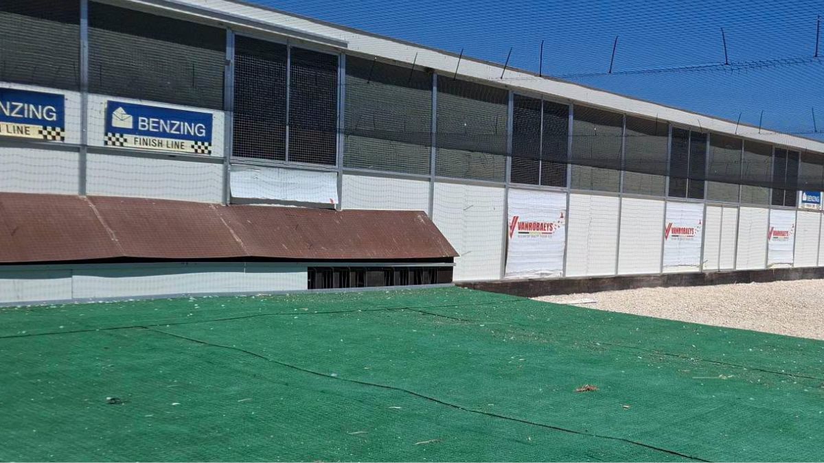 A wide outdoor shot of the long, modern OLR Lasterie-Zwiep racing pigeon loft facility located in Abanilla, Murcia, Spain. The large structure features extensive upper mesh screening for open-air ventilation, clean white lower wall paneling, and a wide green turf landing platform stretched out in the foreground. On the left side of the building, prominent "Benzing Finish Line" electronic timing signs are displayed, with several red "Vanrobaeys" sponsor banners visible along the front facade.