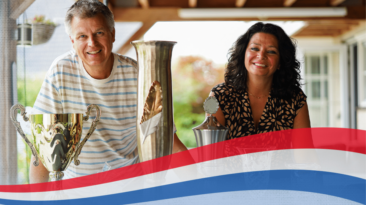 Gerard and Katiuska Schalkwijk smiling behind their national marathon racing pigeon trophies with a red, white, and blue banner overlay.