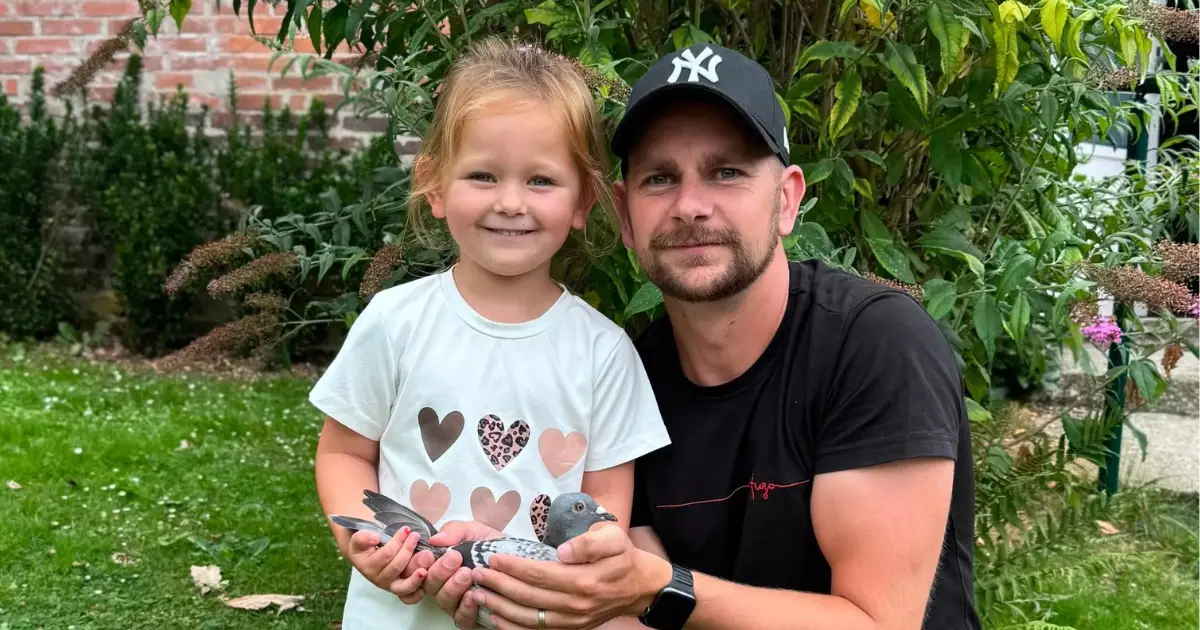 Tim Penders and a young girl holding a racing pigeon together outdoors in a green garden setting.